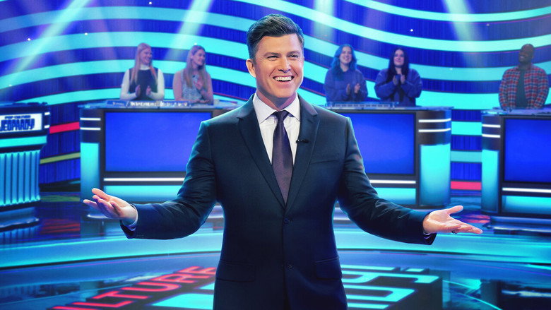 Colin Jost standing at the *Jeopardy!* podium, mid-laugh, as the game board glows behind him