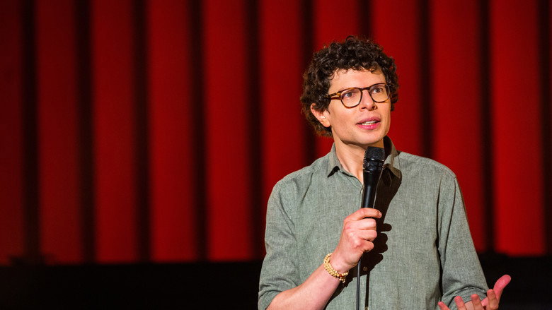 A dimly lit stage, Simon Amstell standing alone under a single spotlight, his posture open but vulnerable, as if inviting the audience into a confession.
